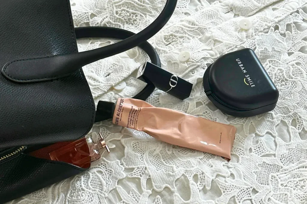Flat lay of everyday essentials including an Urban Smile retainer case, handbag, lipstick, perfume and skincare on lace fabric, showing a retainer as part of a daily routine.